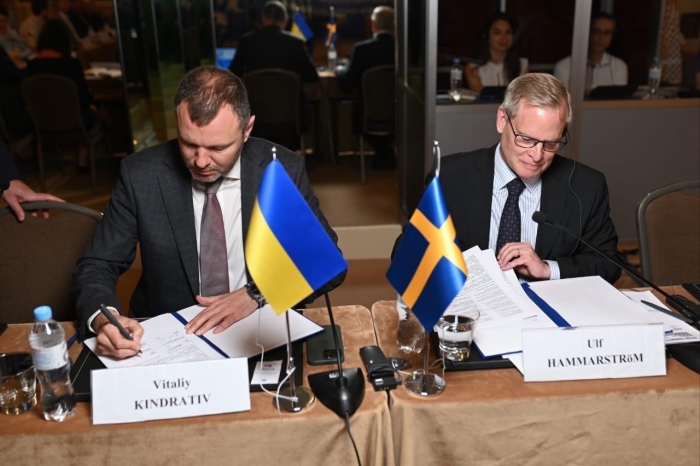 Ulf Hammarström och Vitaliy Kindrativ skriver under var sitt avtal framför en svensk och ukrainsk flagga som står på bordet framför dem.